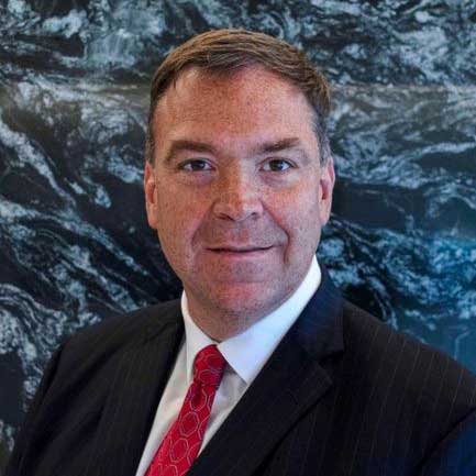 Representative Turco is dressed in a suit and smiling, and standing in front of a stone wall.